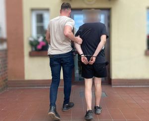 Funkcjonariusz Wydziału Kryminalnego prowadzi mężczyznę w kajdankach. Funkcjonariusz ubrany jest po cywilnemu.