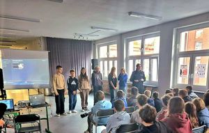 Prelekcja policjantki w szkole. Na korytarzu na krzesełkach siedzi młodzież. Funkcjonariuszka przeprowadza prelekcję.