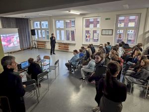 Prelekcja policjantki w szkole. Na korytarzu na krzesełkach siedzi młodzież. Funkcjonariuszka przeprowadza prelekcję.