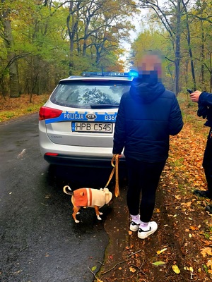 Na zdjęciu stoi kobieta z psem, która zgubiła się w lesie. W tle znajduje się radiowóz policyjny